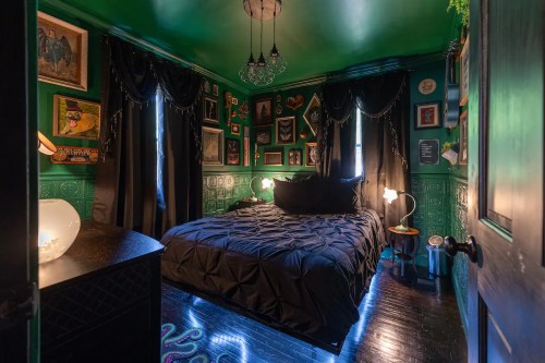 Cozy bedroom with green walls, black bedding, dark curtains, and eclectic wall art, illuminated by soft lighting.