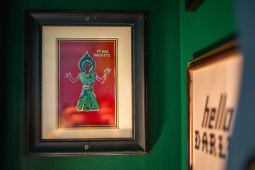 A framed drawing of a green figure with a hood, captioned "It has pockets," against a dark green wall.
