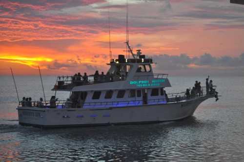 A boat with passengers sails on calm waters at sunset, with vibrant orange and purple skies in the background.