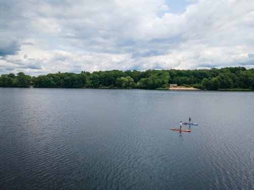 Two people paddleboarding on a calm lake surrounded by trees under a cloudy sky.