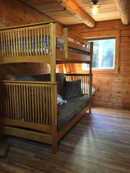 Cozy wooden cabin interior featuring a bunk bed and a sofa by a window, with warm lighting and wooden walls.