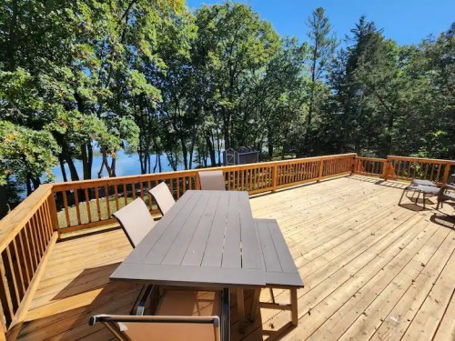A spacious wooden deck with a table and chairs, surrounded by trees and overlooking a serene lake.