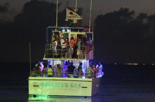 A boat named "Murphy's Law" with people on deck, illuminated by colorful lights, sailing at night.