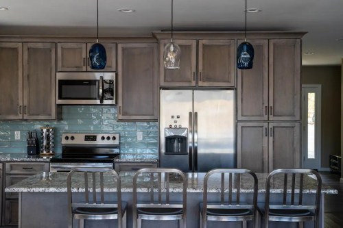Modern kitchen with gray cabinets, stainless steel appliances, and a granite island with bar stools. Blue pendant lights hang above.