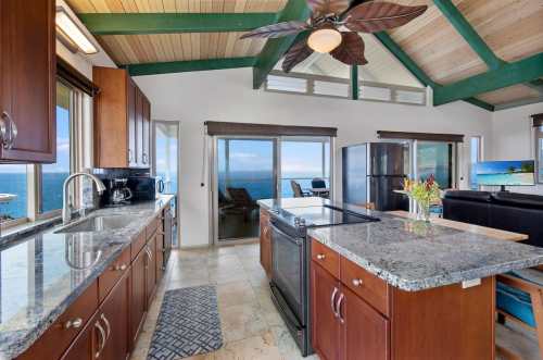 Bright kitchen with granite countertops, wooden cabinets, and ocean views through large windows. Ceiling fan above.