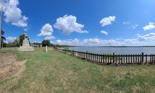 A panoramic view of a river with a grassy area, a fence, and a monument under a blue sky with fluffy clouds.