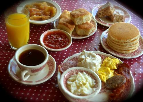 A breakfast spread featuring pancakes, biscuits, eggs, bacon, sausage, grits, fruit preserves, and orange juice.