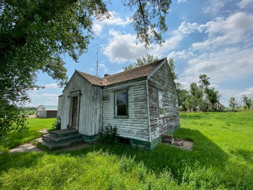 A weathered, small house with a wooden exterior, surrounded by green grass and trees under a partly cloudy sky.