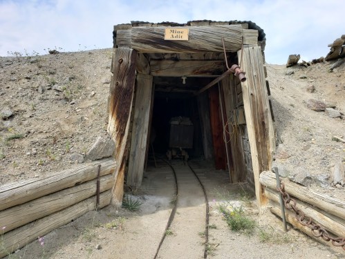 Entrance to an old mine adit with wooden beams and a cart on tracks, surrounded by rocky terrain.