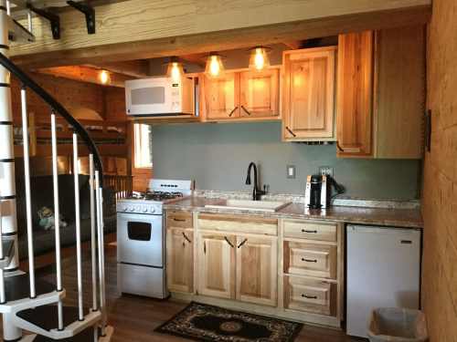 Cozy kitchen with wooden cabinets, stove, microwave, and a spiral staircase in a rustic cabin setting.