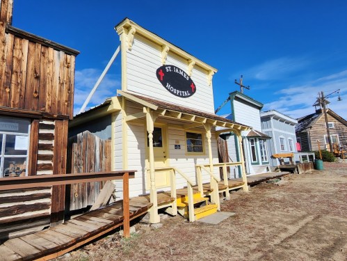 A historic wooden building labeled "St. James Hospital" in a ghost town setting, with blue skies above.
