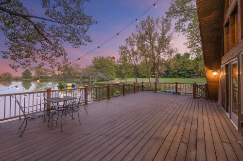 A wooden deck overlooking a calm lake at sunset, with string lights and trees in the background.