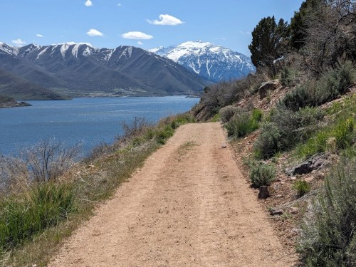 A dirt path along a lake, with mountains and snow-capped peaks in the background under a clear blue sky.