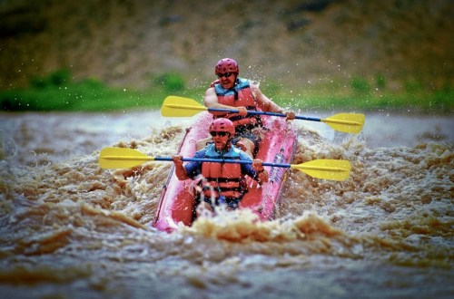 Two people in a pink kayak navigate through turbulent brown waters, wearing helmets and paddling with yellow oars.