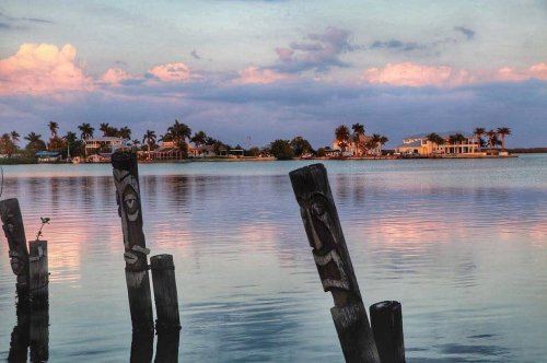 Sunset over calm water with wooden posts in the foreground and palm trees and houses on the distant shore.