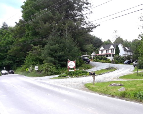 A winding gravel driveway leads to a house surrounded by trees, with parked cars and a sign reading "One More Wonder."