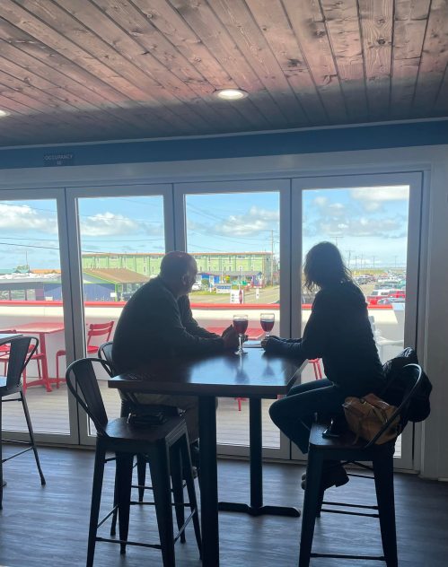 Two people sit at a table by a large window, enjoying drinks and looking out at a scenic view.