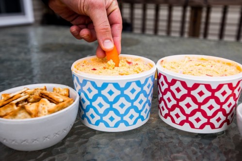 A hand dips a carrot stick into two colorful bowls of dip, with a small bowl of crackers nearby.