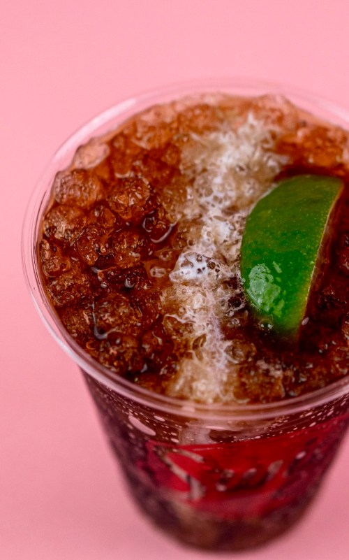 A close-up of a fizzy cola drink with ice and a slice of lime in a clear cup against a pink background.
