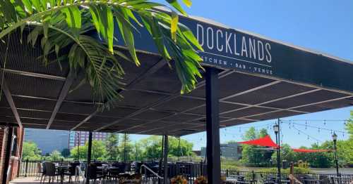 Outdoor seating area of Docklands Kitchen Bar Venue, featuring tables, greenery, and a clear blue sky.