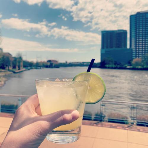 A hand holds a refreshing cocktail with a lime slice, overlooking a river and city skyline under a bright sky.