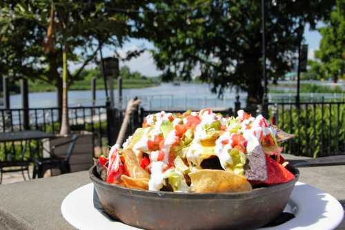 A vibrant plate of nachos topped with lettuce, tomatoes, and sour cream, set against a scenic riverside backdrop.