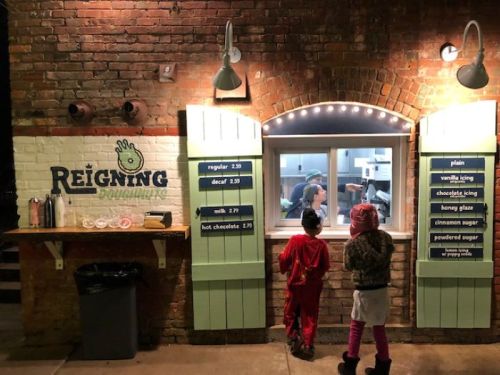 Two children in winter attire stand at a brightly lit food window labeled "Reigning Donuts," looking at the menu.