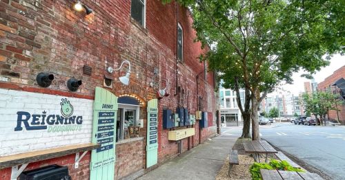 A brick building with a donut shop sign, outdoor seating, and trees lining the sidewalk in an urban setting.