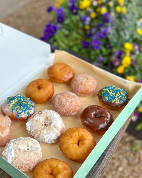 A box of assorted donuts, including glazed, powdered, and frosted, with colorful sprinkles, set against a floral background.
