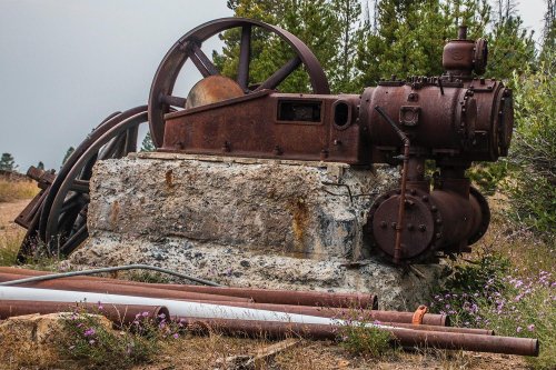 Rusty vintage machinery on a stone base, surrounded by overgrown grass and wildflowers, with trees in the background.