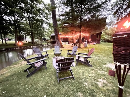 A cozy outdoor setting with wooden chairs arranged around a fire pit near a pond, surrounded by trees and soft lighting.