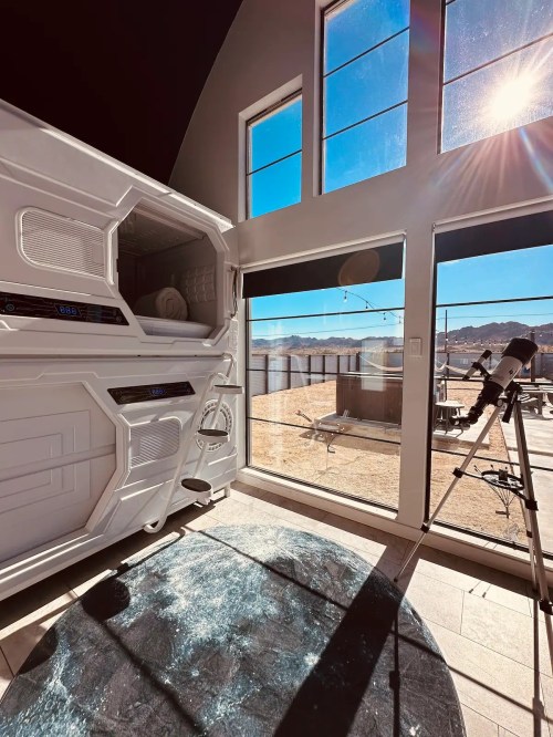A modern room with large windows, a futuristic pod, and a telescope, overlooking a sunny outdoor landscape.
