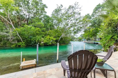 A serene view of a river surrounded by lush greenery, with wooden chairs and a small dock in the foreground.