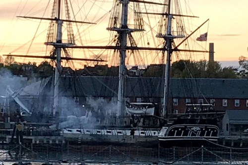 A historic ship docked at sunset, with smoke rising and an American flag visible in the background.