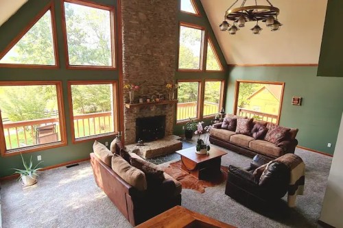 Cozy living room with large windows, stone fireplace, and comfortable seating, surrounded by greenery.
