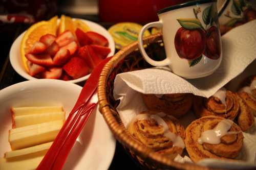 A basket of cinnamon rolls with icing, a cup, and plates of sliced apples, strawberries, and orange wedges.