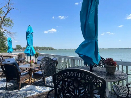 A scenic lakeside patio with blue umbrellas, chairs, and tables, overlooking calm water and a clear blue sky.