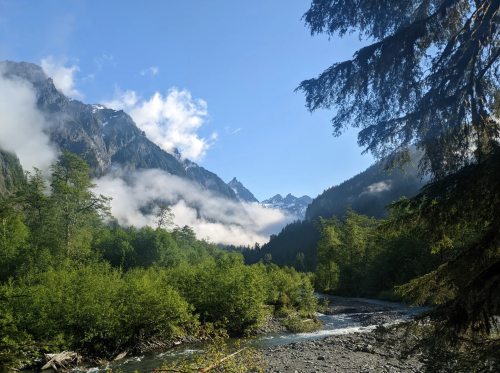 A serene mountain landscape with lush greenery, a river, and misty peaks under a clear blue sky.