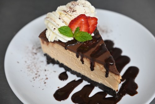 A slice of chocolate cheesecake topped with whipped cream, a strawberry, and mint, drizzled with chocolate sauce.