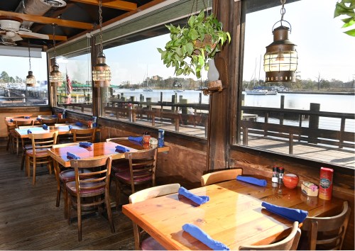 Cozy restaurant interior with wooden tables, blue napkins, and large windows overlooking a waterfront view.
