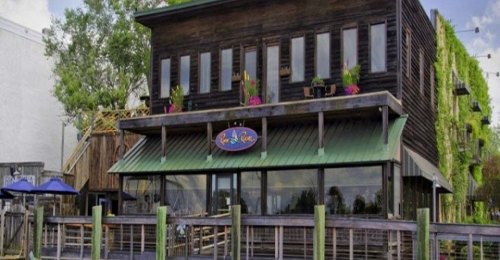 A rustic waterfront building with a green roof, large windows, and colorful flowers, surrounded by wooden posts.