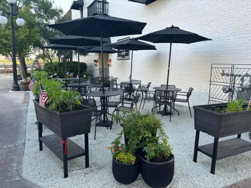 Outdoor seating area with black umbrellas, tables, and planters, featuring greenery and small American flags.