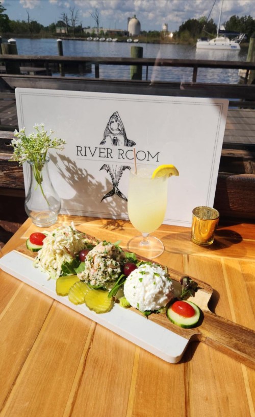 A wooden platter with three seafood salads, pickles, and cherry tomatoes, alongside a drink and a sign for "River Room."