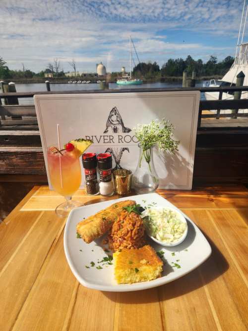 A plate of fried fish, rice, coleslaw, and cornbread, with a cocktail and condiments, by a scenic waterfront view.
