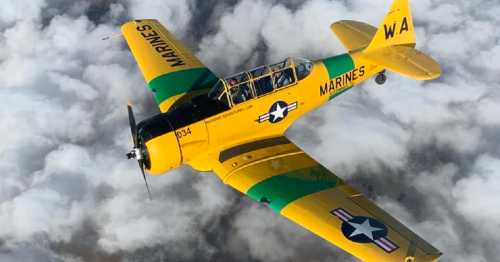 A yellow and green military training aircraft flying above fluffy white clouds.