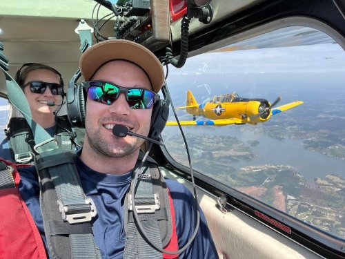 A pilot and passenger in a small aircraft, smiling, with a vintage yellow plane flying nearby against a scenic backdrop.