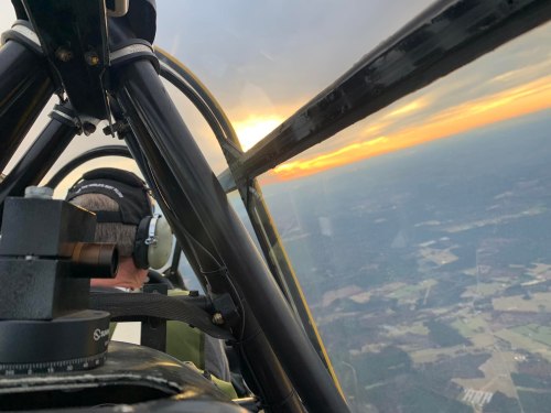 Aerial view from a cockpit at sunset, showing a pilot and landscape below.