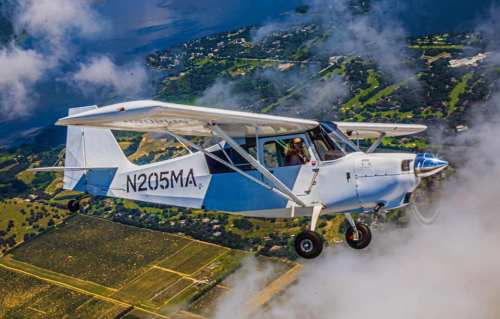 A small white aircraft flying above clouds with a scenic landscape and water below.