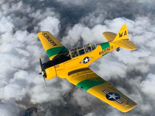 A yellow and green military training aircraft flying above fluffy white clouds.