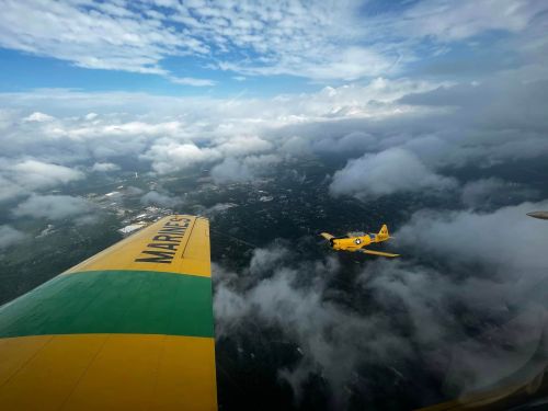 A yellow airplane flying alongside a green and yellow wing above clouds, with a blue sky in the background.